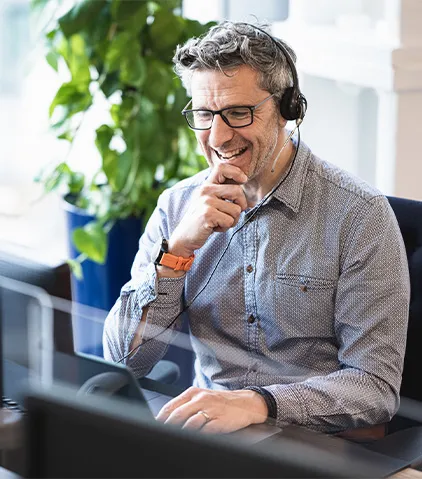 Person trägt ein Headset und arbeitet an einem Schreibtisch mit Computer in einer modernen Büroumgebung mit Pflanzen im Hintergrund.