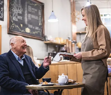 Gast bezahlt mit Karte bei Bedienung in gemütlichem Café mit Holzeinrichtung und Kreidetafel.