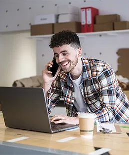 Person nutzt ein Smartphone, während sie an einem Laptop an einem Schreibtisch mit Kaffeebecher und Büromaterial im Hintergrund arbeitet.