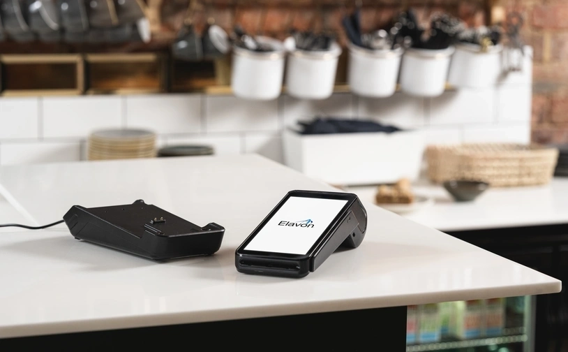 Zahlungsterminal mit Elavon-Logo auf einer weißen Arbeitsplatte in einem Café mit Tassen und Regalen im Hintergrund