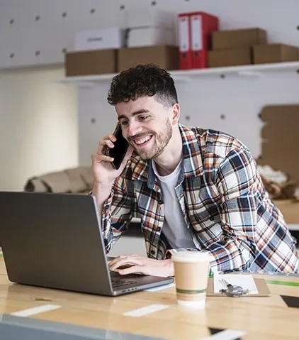 Person telefoniert mit einem handy und arbeitet an einem laptop an einem schreibtisch mit einem kaffeebecher und dokumenten