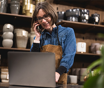  Person mit Schürze telefoniert mit dem Handy und benutzt einen Laptop in einem Geschäft mit Regalen voller Töpfe