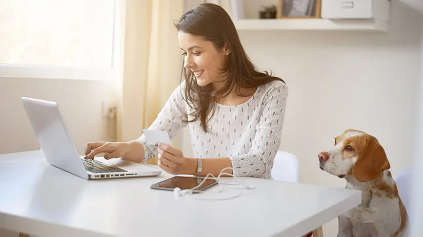 Person sitzt an einem tisch und nutzt einen laptop hält einen beleg mit einem smartphone und kopfhörern auf dem tisch und ein hund sitzt daneben