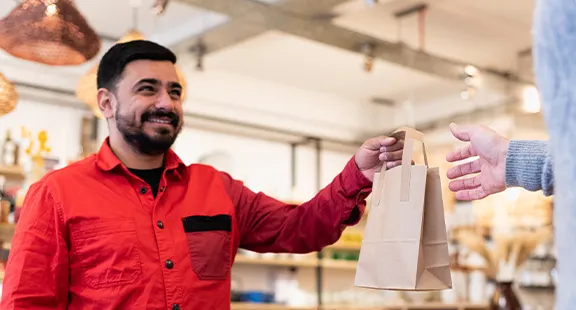 Person im roten shirt übergibt papiertüte an kunden in geschäft mit regalen im hintergrund