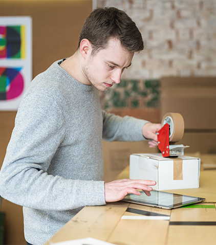 Eine Person in einem hellgrauen Pullover verwendet einen Klebebandabroller, um einen Karton auf einem Holztisch zu versiegeln, mit weiteren Kartons und bunter Kunst im Hintergrund.